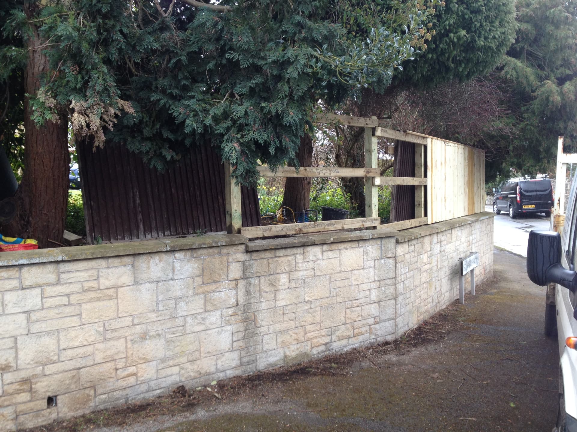 Feather-edge fencing being installed on top of a stone retaining wall beneath overhanging trees